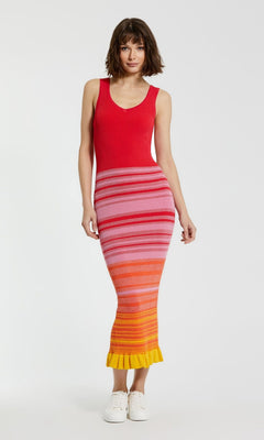 A woman stands against a plain white background wearing the Mac Duggal Red Casual Wedding-Guest Dress K12007, a striped tank dress in red, pink, orange, and yellow hues, paired with white sneakers.