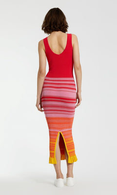 A person stands facing away, wearing the Mac Duggal Red Casual Wedding-Guest Dress K12007— a sleeveless striped tank dress with gradient pink, orange, and yellow stripes, a back slit with yellow trim—paired with white shoes against a plain background.