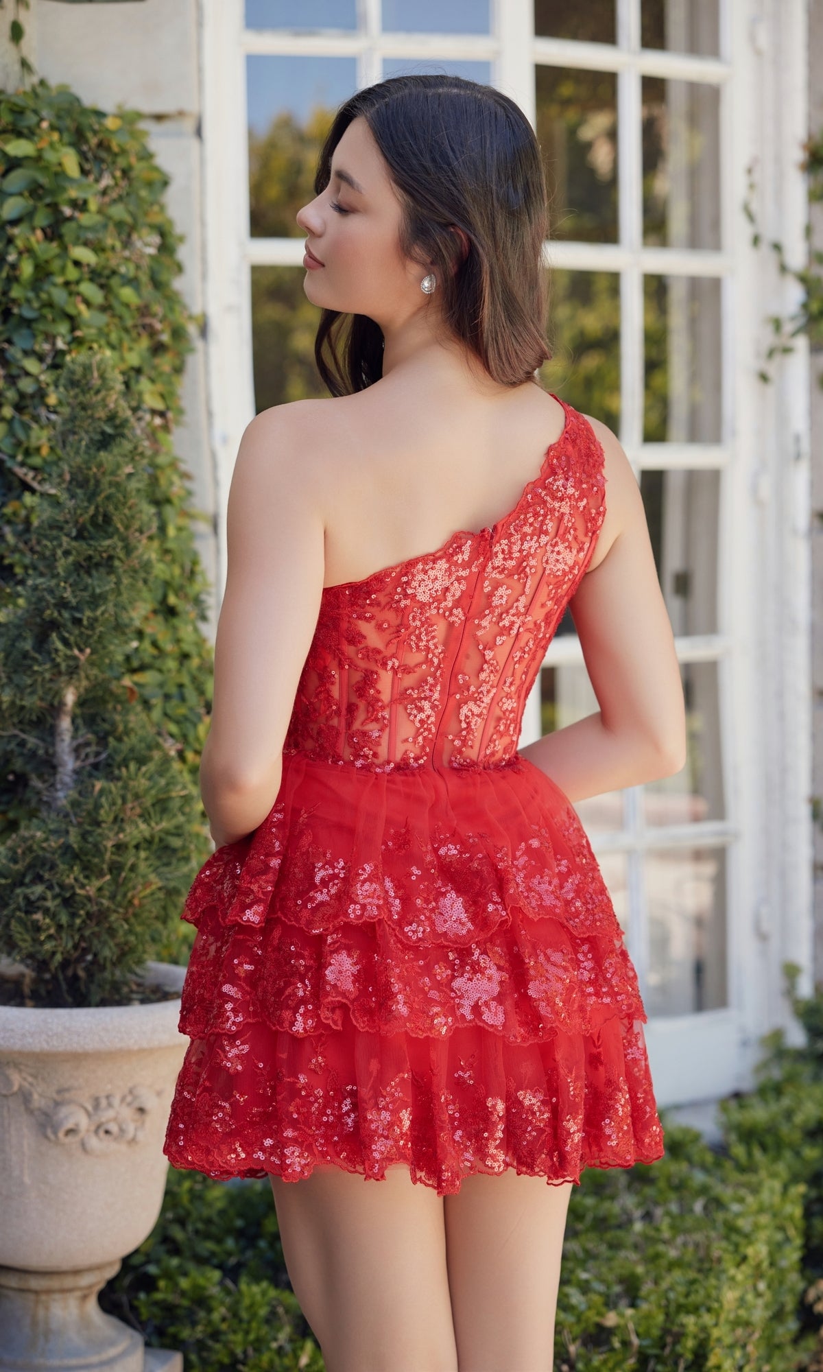 A woman stands outdoors in front of a window, facing away from the camera, wearing the One-Shoulder Short Sequin Homecoming Dress K855 in red with lace and tiered tulle ruffles.