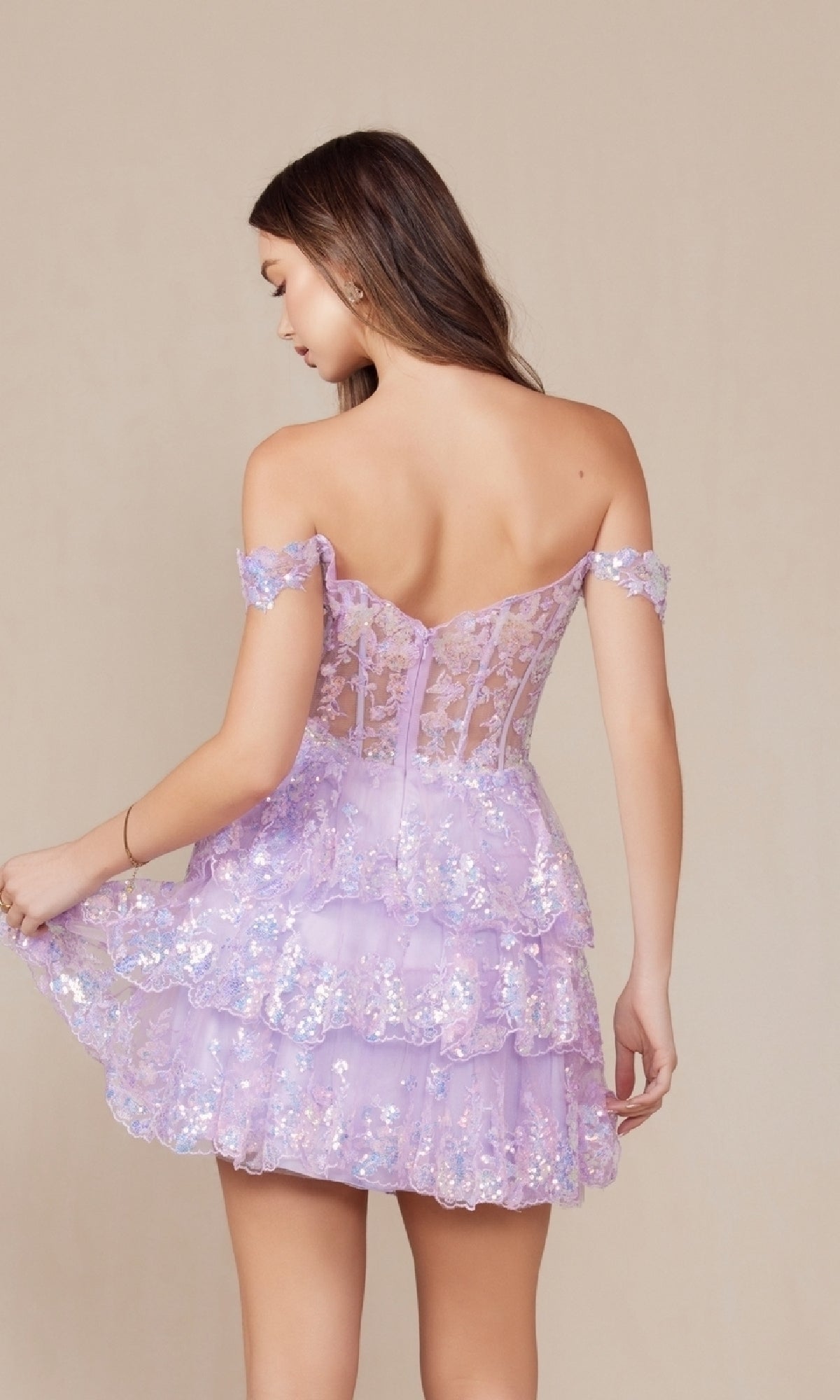 A woman faces away wearing the Off-Shoulder Short Ruffled Sequin Hoco Dress K856, a lilac gown featuring floral lace, ruffles, and sequin details, while holding one side of its layered skirt.