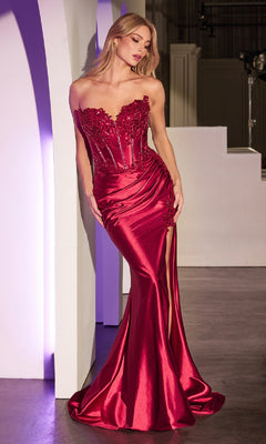A woman wearing the Ladivine KV1116 Strapless Long Corset Prom Dress, with beaded-lace applique bodice and a skirt slit, stands indoors before a white geometric backdrop.