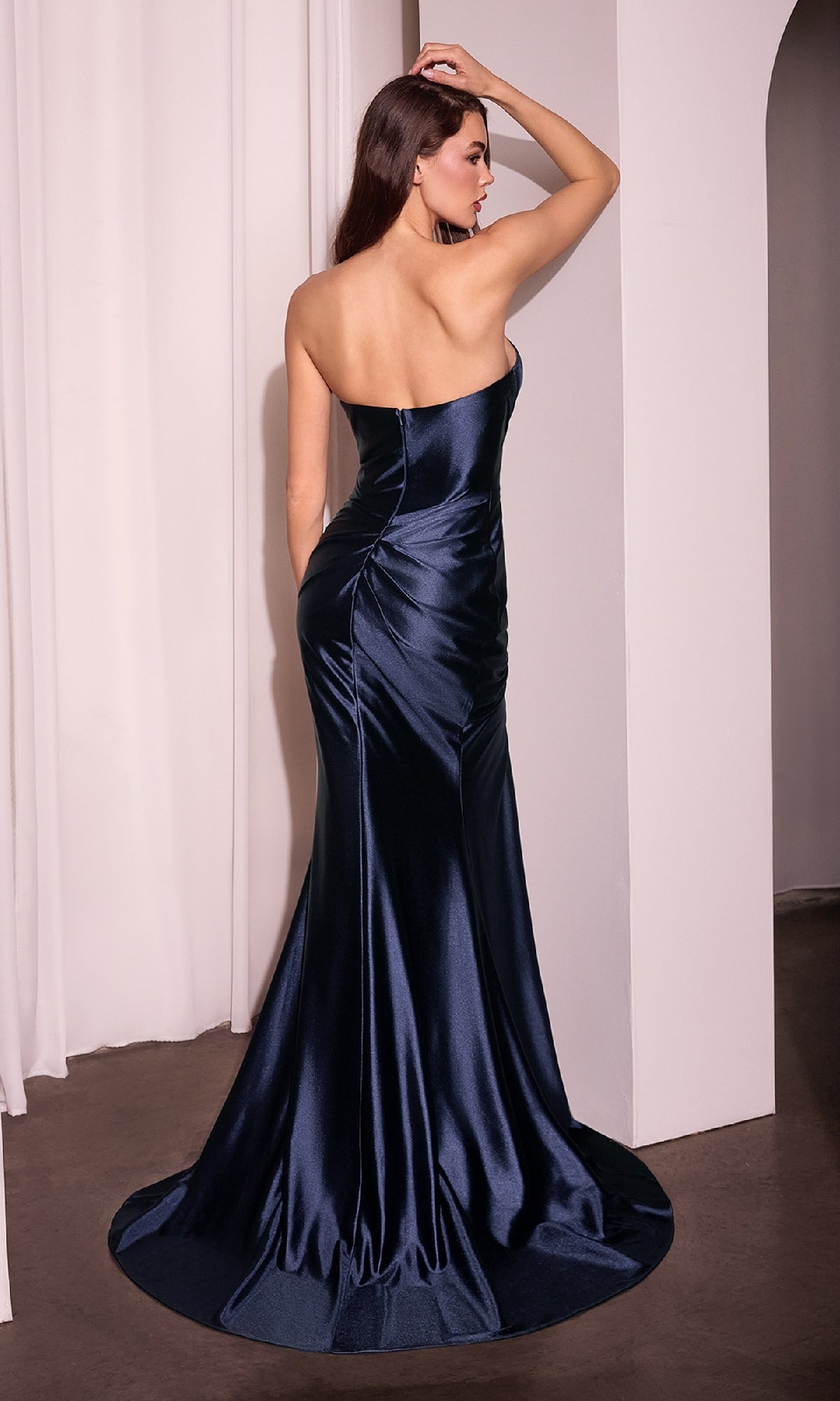 A woman in a dark blue Strapless Long Satin Prom Dress: Ladivine KV1118 stands with her back to the camera, one arm raised and touching her head.