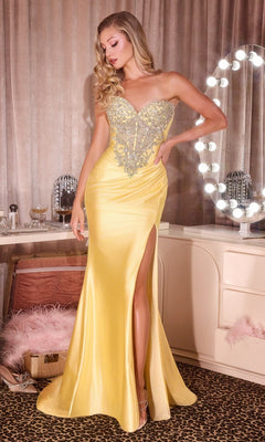 A woman in a yellow Ladivine KV1142 long prom dress with a beaded bodice and high slit stands in a decorated dressing room with a lit vanity mirror.