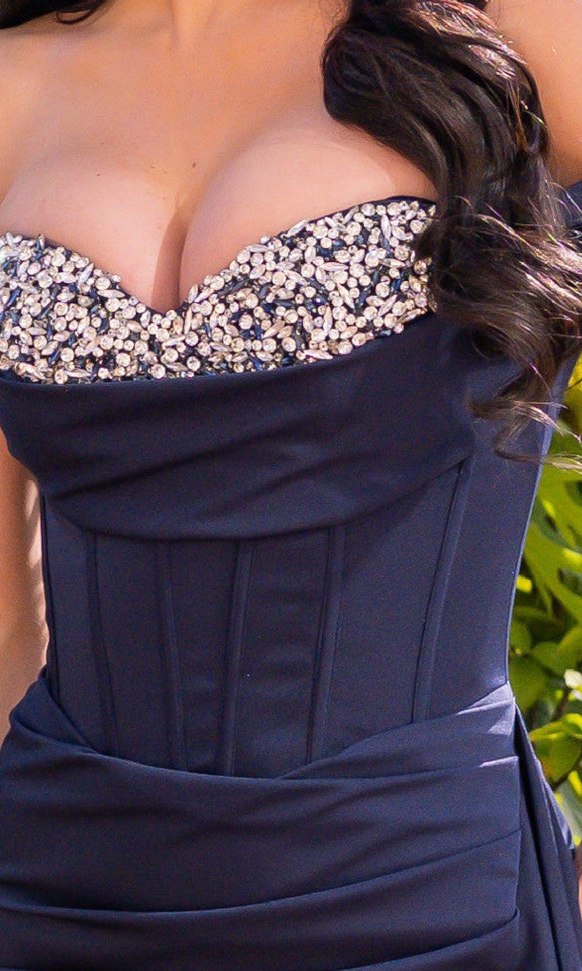 A woman with long, dark wavy hair wears the Off-the-Shoulder Corset Prom Dress: Katrina by Velvi in navy blue, featuring ruching and a jeweled neckline, shown from shoulders to waist.