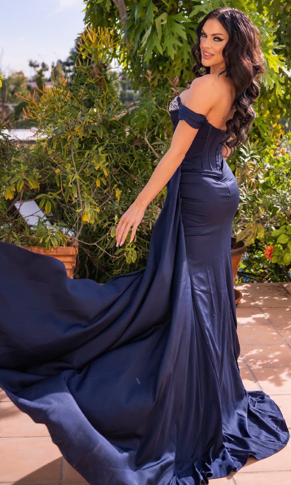 Woman in the Off-the-Shoulder Corset Prom Dress: Katrina by Velvi poses on a patio, holding out her fitted navy blue gown with lush greenery in the background.
