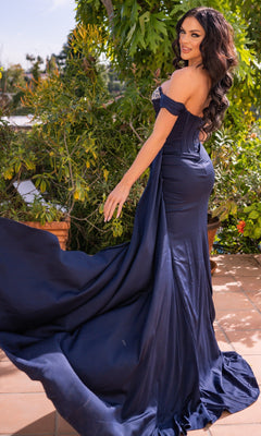 Woman in the Off-the-Shoulder Corset Prom Dress: Katrina by Velvi poses on a patio, holding out her fitted navy blue gown with lush greenery in the background.