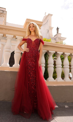 A woman stands outdoors in front of white railings, wearing the Overskirt Long Sequin Prom Dress: Nox Anabel L1362—an elegant red off-the-shoulder gown with floral embellishments, tulle details, and a floor-length silhouette.