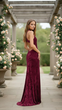 A woman in the High-Neck Velvet Prom Gown: La Femme 25783 stands on a stone path, surrounded by rose bushes and pergola columns, glancing back over her shoulder.