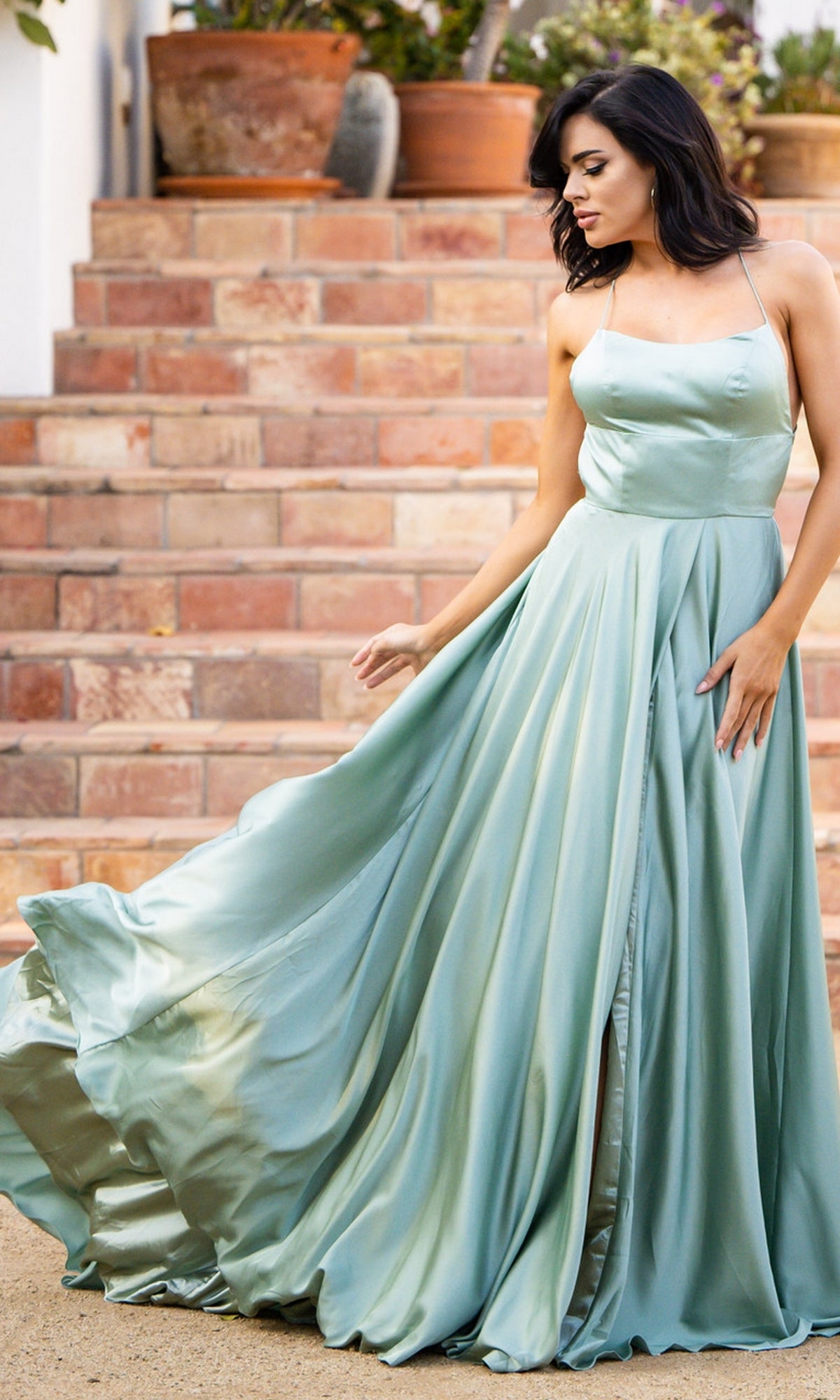 A woman wearing the Velvi Long Satin Prom Dress Liliana stands outdoors, holding out one side of her flowing skirt. Brick steps and potted plants are visible in the background.