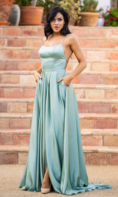 A woman stands outdoors on stone steps in the Velvi Long Satin Prom Dress Liliana, light blue with spaghetti straps, hands in pockets and beige high heels visible.