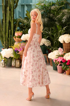 A woman wearing the Cream Floral Print Casual Midi Dress: Liora and high heels stands indoors, looking over her shoulder, surrounded by potted plants and flowers.