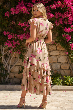 A woman wearing the Lucy Taupe Pink Floral Midi Wedding-Guest Dress stands outdoors in tan heels, ruffles flowing, with pink flowers and a stone wall behind her.