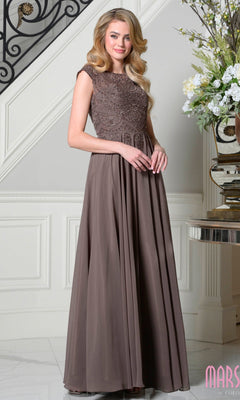 A woman poses indoors near a floral display and decorative staircase, wearing the Marsoni M238 long formal dress in dark taupe—a sleeveless, floor-length gown with an embellished lace bodice and flowing skirt.
