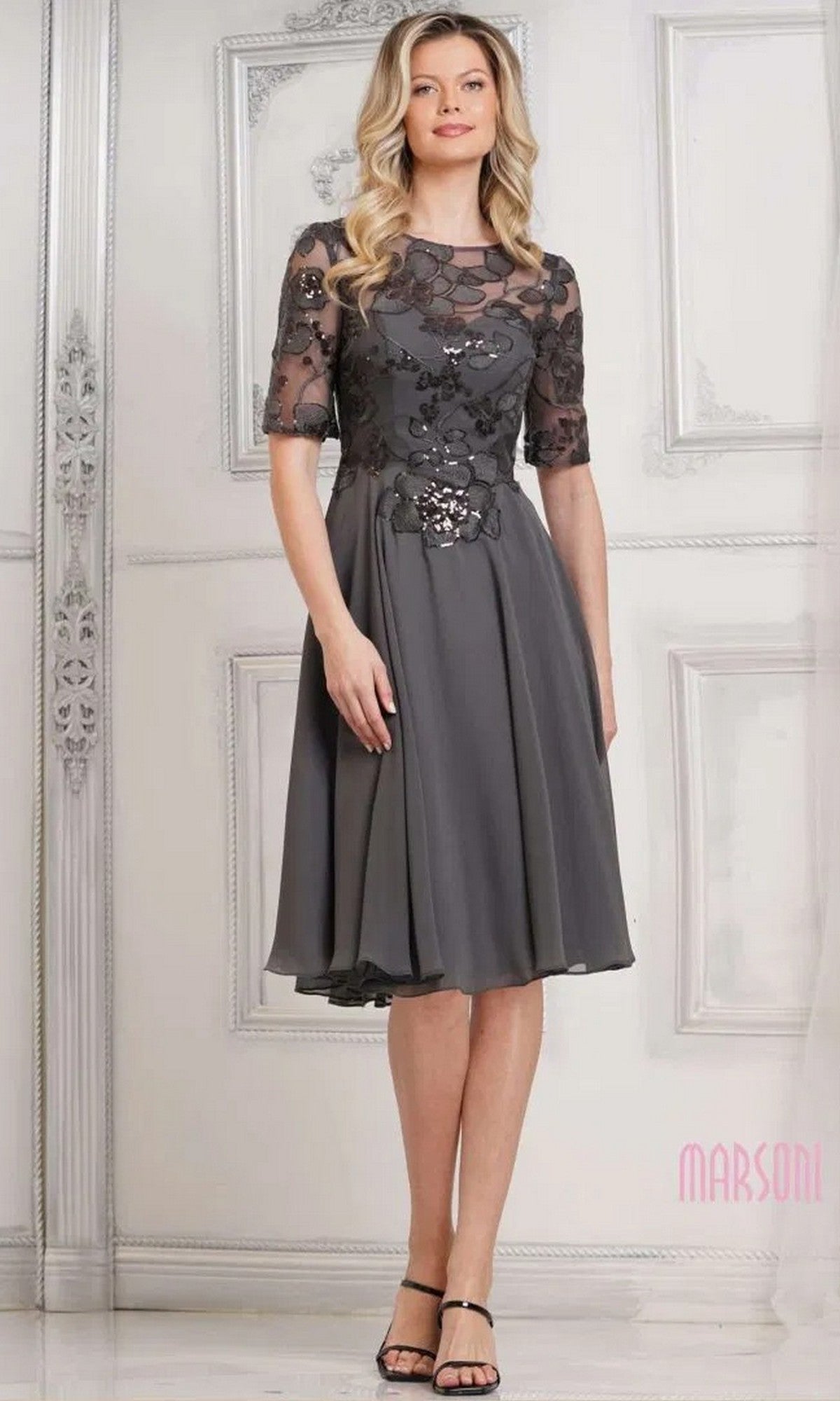 A woman stands in an ornate room wearing the Marsoni M286S semi-formal knee-length dress in charcoal, featuring floral embroidery, sheer sleeves, an a-line chiffon skirt, and black heeled sandals.