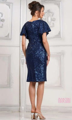 A woman stands indoors, facing away, wearing the Marsoni M318S knee-length semi-formal dress in navy blue sequins with beige high-heeled sandals against a white ornate wall.