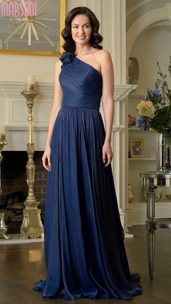 A woman in the navy Marsoni M332 long formal dress, featuring a floor-length, one-shoulder design with 3D floral embellishments, stands indoors by a fireplace and decorative shelves.