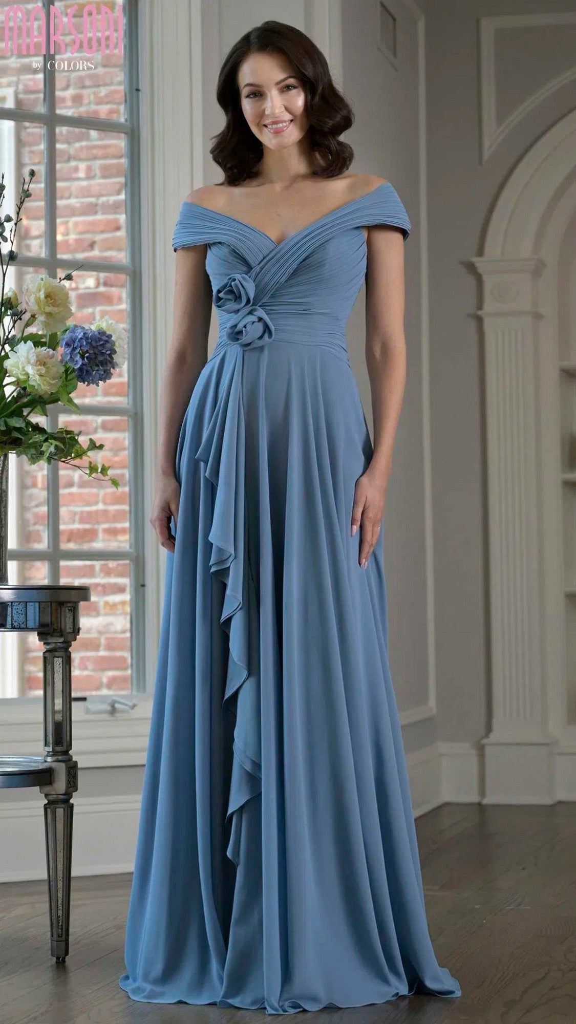 A woman in a Marsoni M333 long formal dress—a floor-length, dusty blue gown—stands indoors near a window and a floral arrangement.