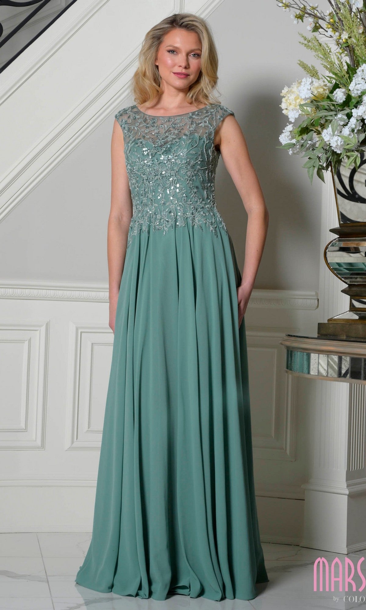 A woman stands indoors wearing the Marsoni M340—a sage, cap-sleeve long A-line formal dress with a beaded bodice and flowing skirt, perfect for those seeking an elegant look.