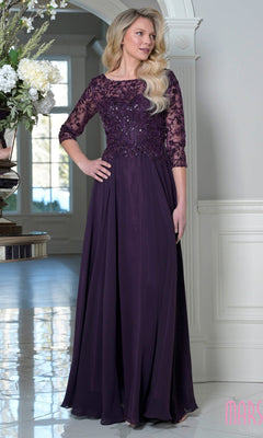 A woman stands indoors near a large window and floral arrangement, wearing the Marsoni M340SL long formal dress in eggplant with a modest beaded-embroidered bodice and A-line chiffon skirt.
