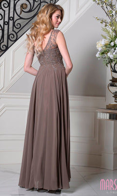 A woman with long wavy hair wears the Marsoni M340 Cap-Sleeve Long A-Line Formal Dress in taupe, featuring floral lace detailing on the sheer back, standing beside a staircase and an elegant floral arrangement.