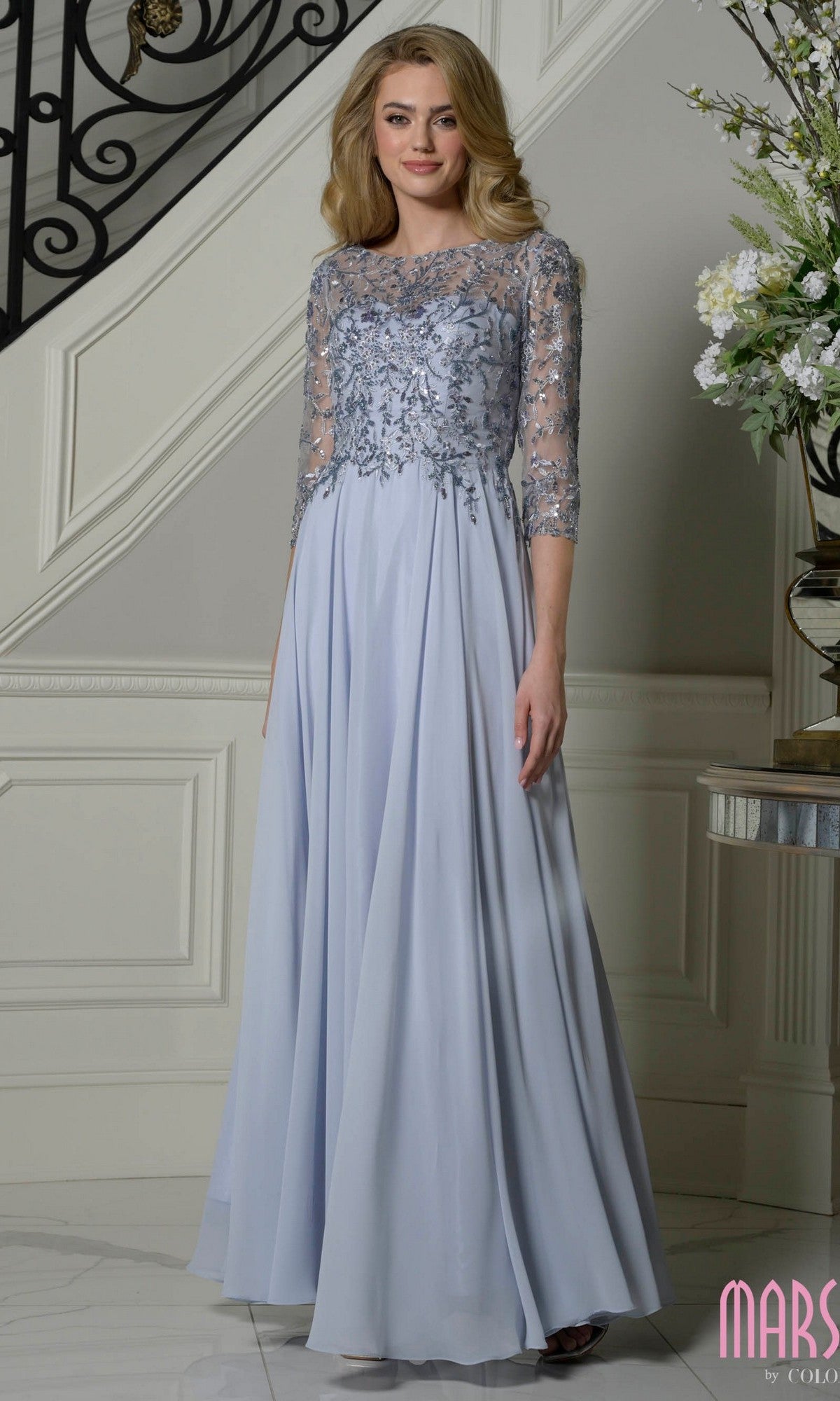 A woman stands indoors in a dusty blue Marsoni M340 Cap-Sleeve Long A-Line Formal Dress, featuring a lace-embroidered bodice and sheer sleeves, next to a staircase and floral arrangement.