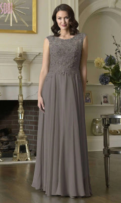 A woman stands indoors in front of a decorated mantel, wearing the Marsoni M340 long formal dress in dark taupe with a lace-embroidered bodice and sleeveless chiffon A-line skirt.