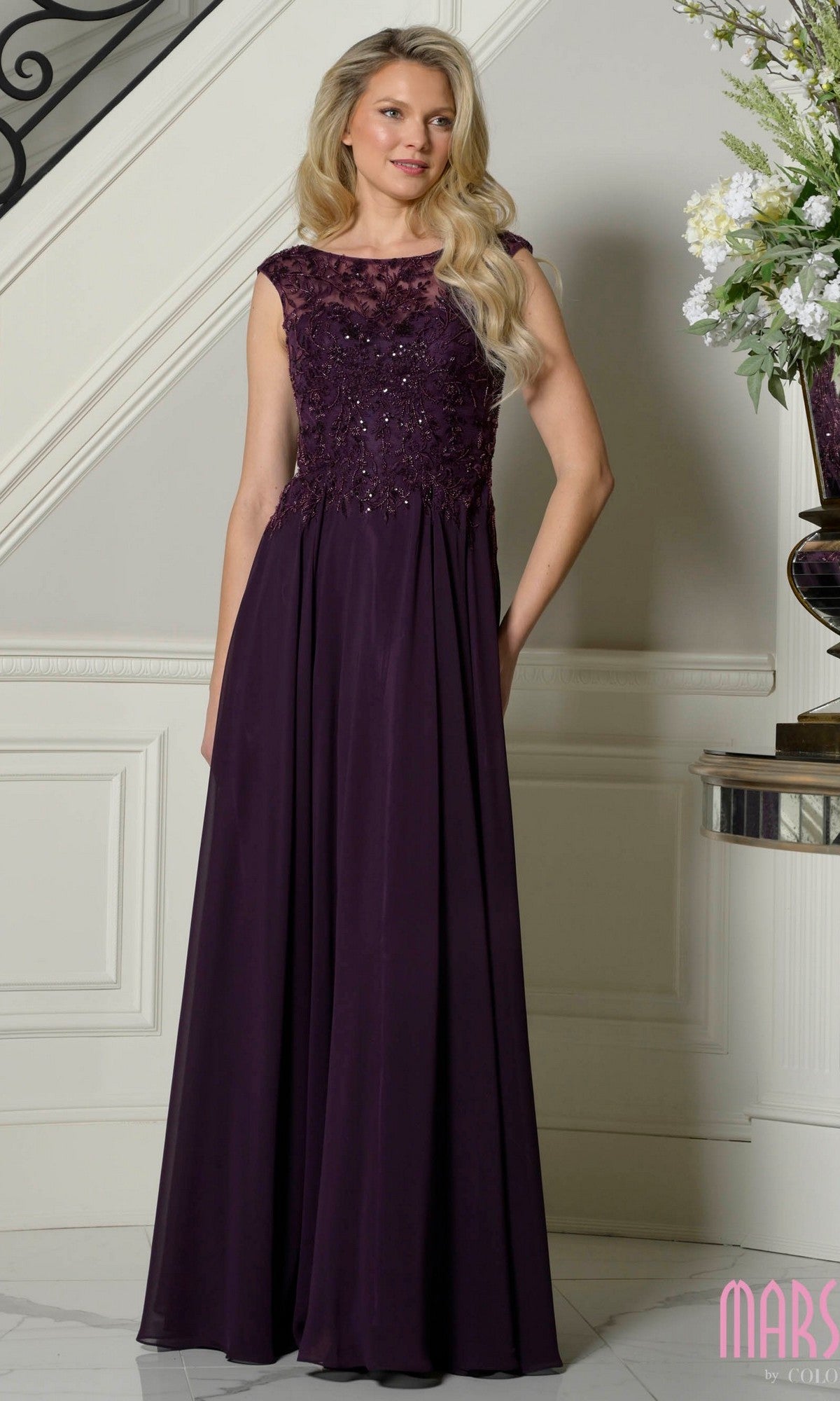 A woman in an eggplant Marsoni M340 cap-sleeve long A-line formal dress with lace and sequin details stands indoors near a staircase and floral arrangement.