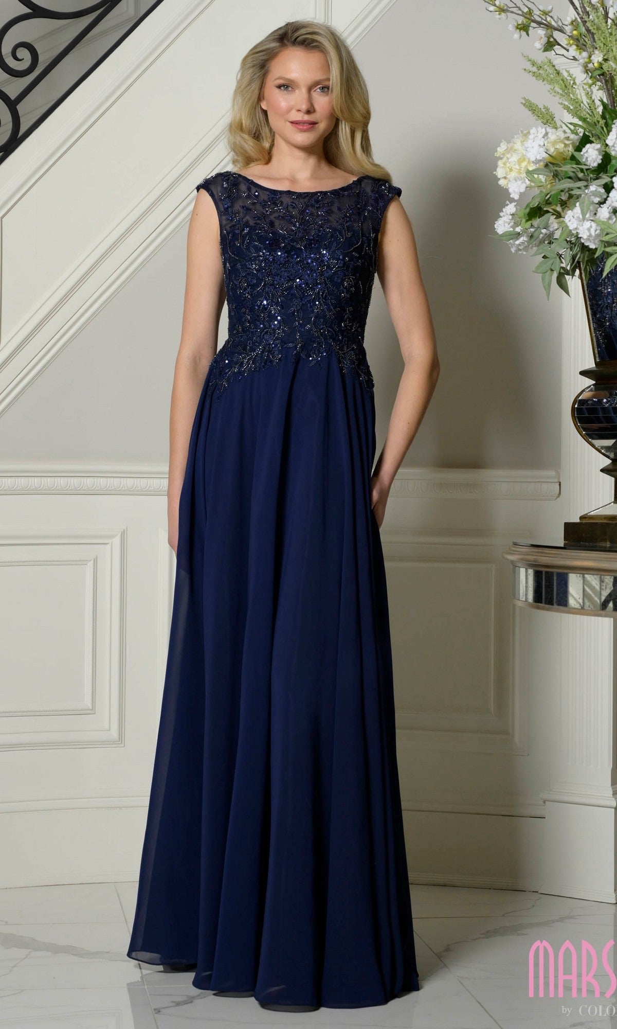 A woman stands indoors wearing the Marsoni M340, a navy cap-sleeve long A-line formal dress with a lace-embroidered bodice.