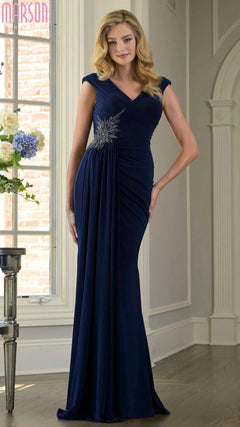 A woman stands indoors wearing the Marsoni M343 Long Formal Dress in navy, featuring a floor-length design with a ruched fitted bodice, cap sleeves, and a silver embellishment at the waist.