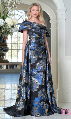 A woman stands indoors in the Marsoni M348 long formal dress, featuring an off-the-shoulder design and floor-length silhouette with royal multi-colored floral jacquard patterns on a black background.