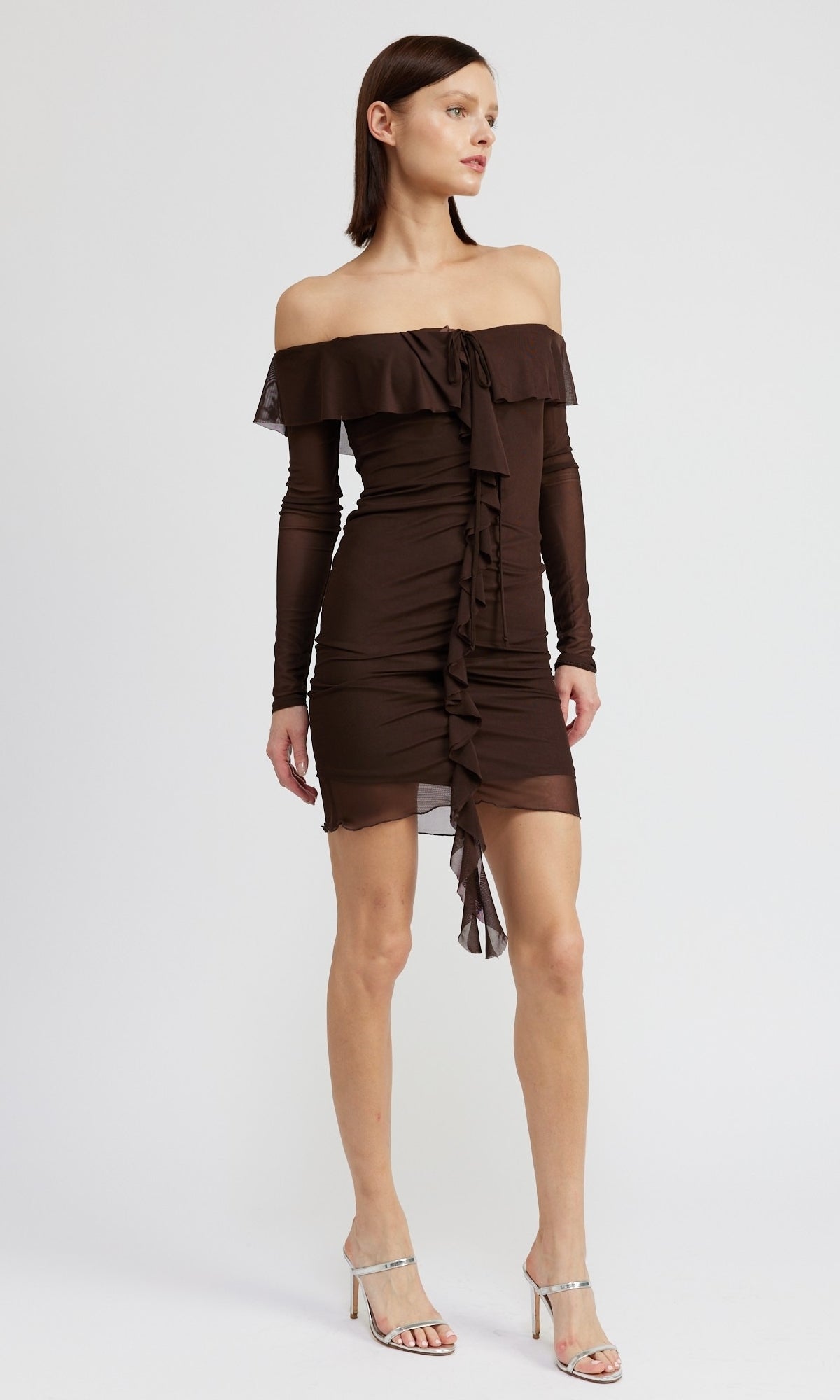 A woman models the Chocolate Brown Short Party Dress with Sleeves, featuring an off-the-shoulder design and ruffle details, paired with silver high-heeled sandals against a plain white background.