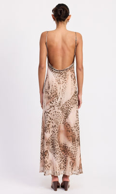 A person stands facing away, wearing the Brown Leopard Cowl-Neck Slip Dress with a sleeveless, open-back design and heeled sandals.