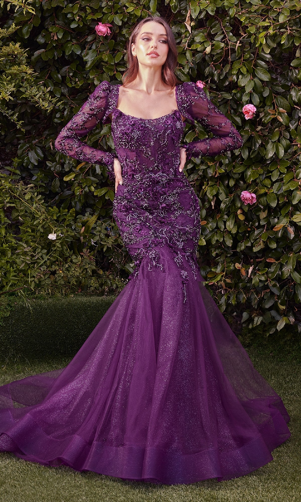 A woman stands outdoors amid greenery and flowers wearing the Andrea & Leo MA101, a long sleeve formal dress with beaded lace, sheer sleeves, and a flared tulle skirt.