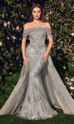 A woman stands outdoors before greenery, wearing the Andrea & Leo MA103 Long Silver Prom Ball Gown with an off-the-shoulder neckline, fitted bodice, and flowing train.