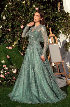 A woman in the Andrea & Leo MA104 Long Sleeve Long Formal Dress poses outdoors by an easel, surrounded by lush greenery and pink flowers.
