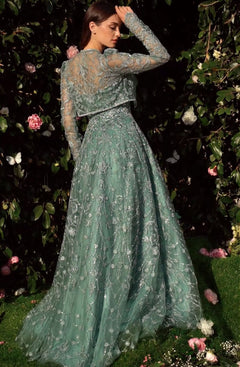 A woman stands in a garden wearing the Andrea & Leo MA104 Long Sleeve Long Formal Dress with embroidered green lace, facing away and touching her head as pink and white flowers bloom in the background foliage.