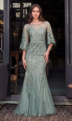 A woman stands in a doorway wearing the Andrea & Leo MA105 Cape-Sleeve Beaded Formal Gown, a floor-length sage green dress featuring intricate beadwork and sheer cape sleeves.