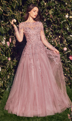 A woman wearing the Andrea & Leo MA108 long sleeve formal gown with lace appliques stands outdoors in front of leafy bushes with pink flowers.