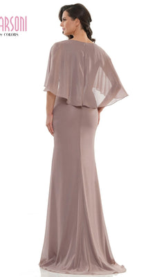 A woman wearing the Marsoni MV1130 long formal dress in mauve, featuring a floor-length design and sheer cape-like overlay, stands facing away from the camera.