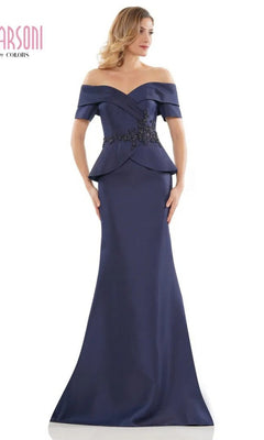 A woman wears the Marsoni MV1144 Long Formal Dress in navy, featuring an off-the-shoulder neckline, fitted bodice, and black floral waist embellishments for an elegant floor-length silhouette.