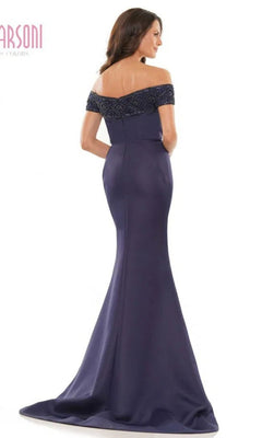 A woman faces away from the camera in the Marsoni MV1184, a long formal dress featuring an off-the-shoulder navy blue design, fitted bodice with beaded details, and an elegant floor-length mermaid skirt.