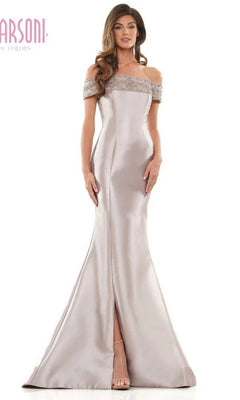 A woman stands against a plain background wearing the Long Formal Dress: Marsoni MV1184 in taupe, featuring a floor-length mermaid skirt with front slit, off-shoulder neckline, and beaded trim.