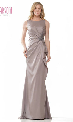 A woman in a sleeveless, floor-length taupe Marsoni MV1233 formal dress with side draping and floral waist embellishment stands against a white background.