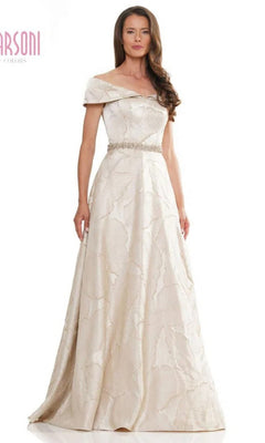 A woman stands against a white background wearing the Marsoni MV1265 long formal dress in champagne, featuring an off-the-shoulder neckline, textured pattern, and jeweled belt for an elegant touch.