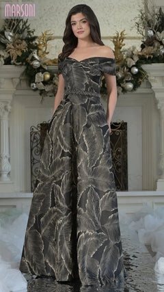 A woman poses indoors in front of a decorated white mantel, wearing the Marsoni MV1265 long formal dress in black—an off-the-shoulder evening gown with a leaf pattern and beaded waistline that exudes timeless elegance.