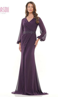 A woman wears the Marsoni MV1273 long formal dress in eggplant, featuring sheer long sleeves and an embellished waist, standing against a plain white background.