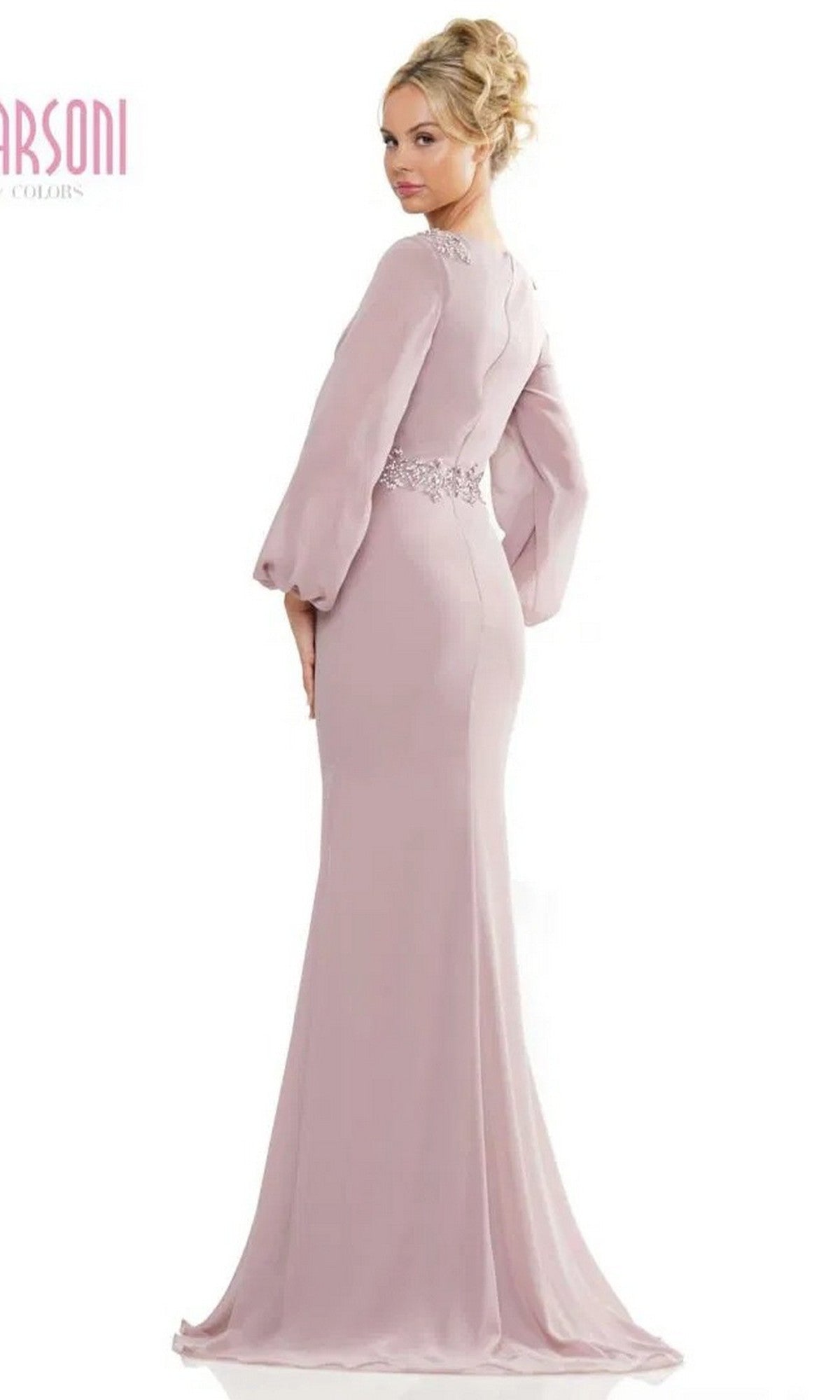A woman wearing the Marsoni MV1273 long formal dress—fitted, floor-length, light mauve with a v-neckline, long sleeves, and lace waist detail—stands facing slightly away from the camera.
