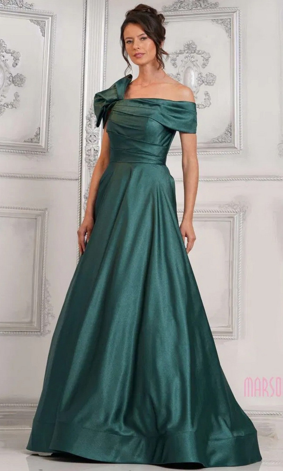 A woman poses elegantly in an ornate room, wearing the Marsoni MV1288 Long Formal Dress in deep green with off-shoulder satin and bow detail.