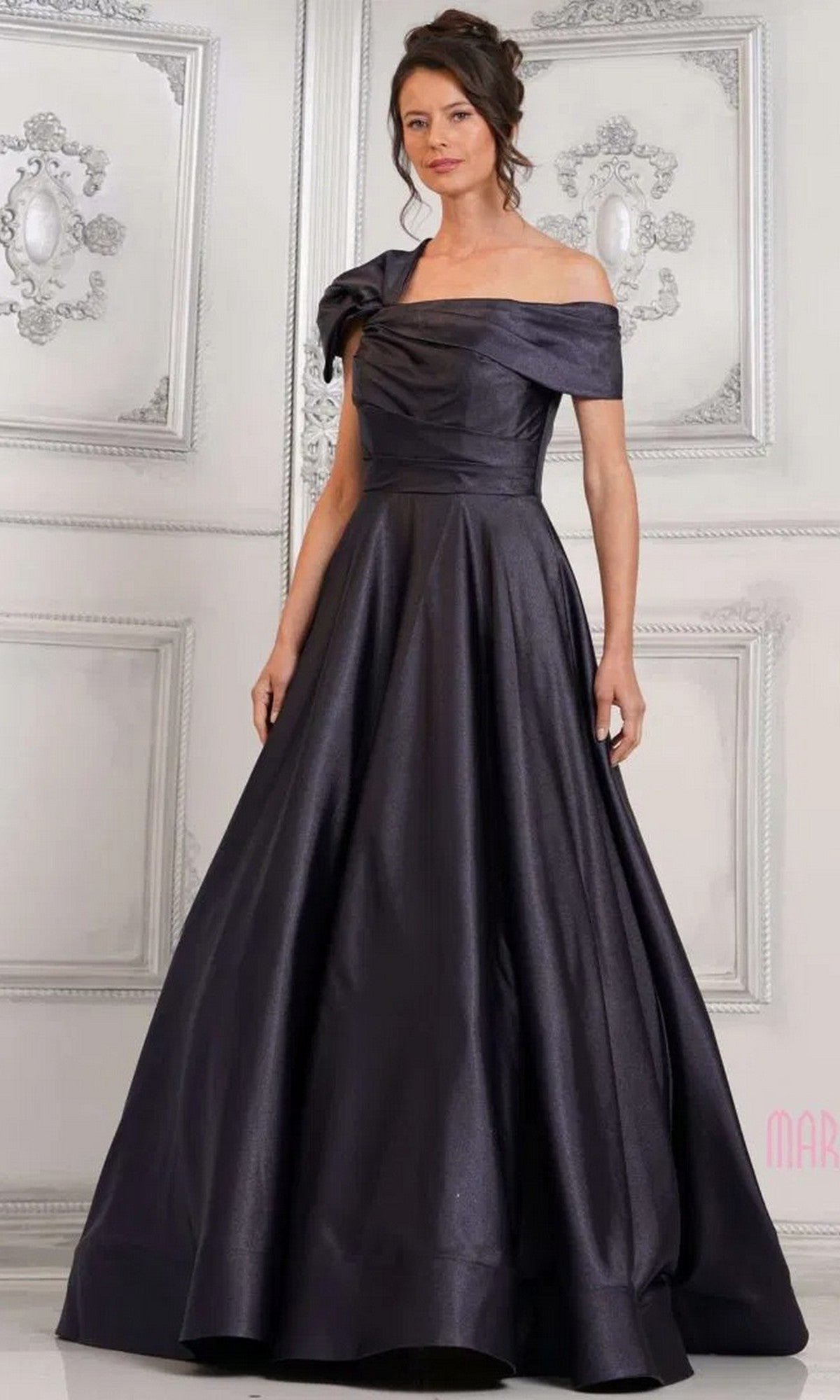 A woman in a navy Marsoni MV1288 long formal dress with an off-shoulder fitted bodice and full skirt stands before an ornate white wall, exuding timeless elegance.