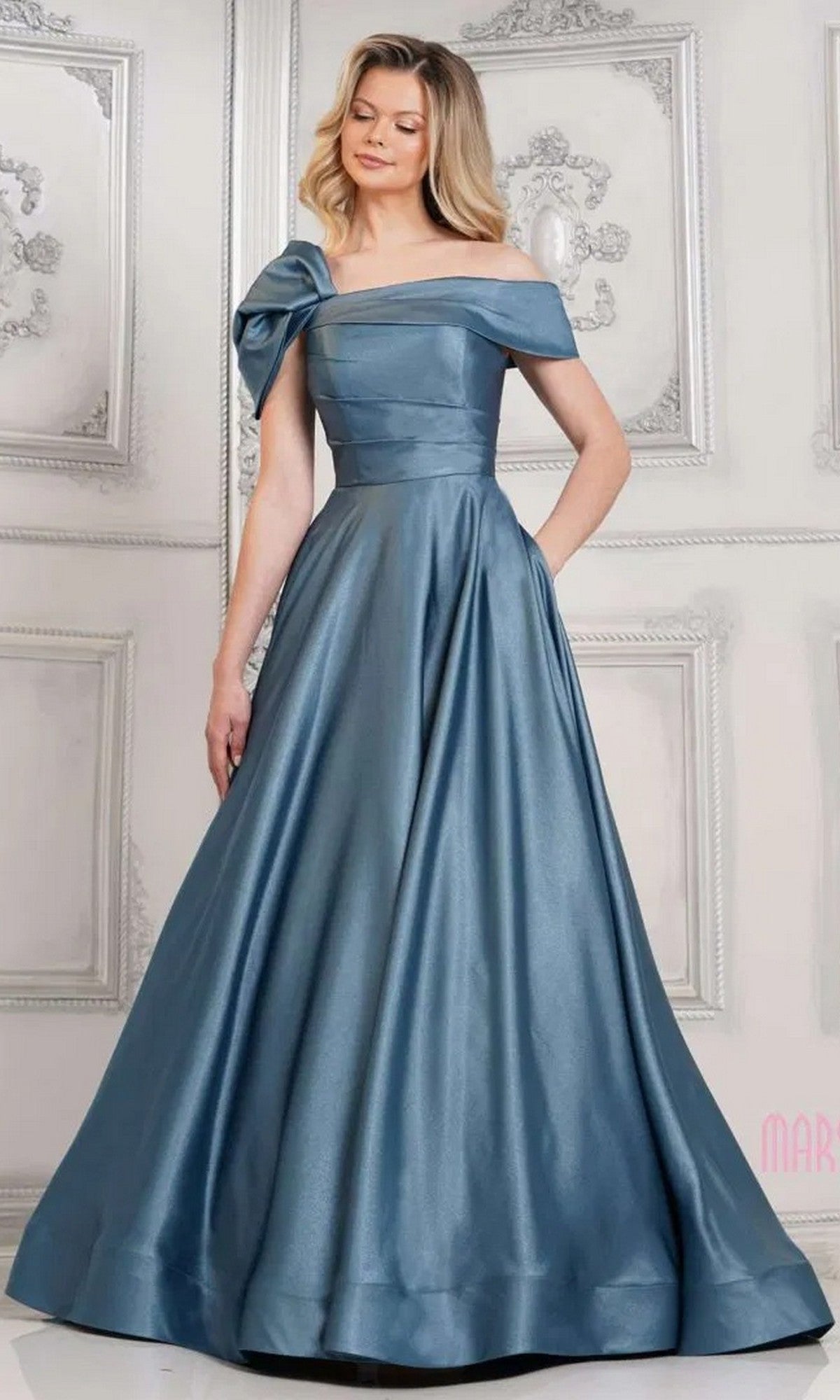 A woman wears the Marsoni MV1288 long formal dress, an off-shoulder blue satin gown with a large shimmery bow on one shoulder, standing gracefully in an elegant, light-colored room.
